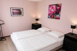 Quarto com duas camas, uma mesa de cabeceira com abajur, um quadro na parede e um candeeiro de chão.