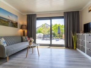 A modern living room with a grey sofa, a small table with a vase, a TV on a wooden cabinet, and a large sliding glass door leading to an outdoor patio. - Comfortable Holiday Apartment in St. Peter Ording (St. Peter-Ording)