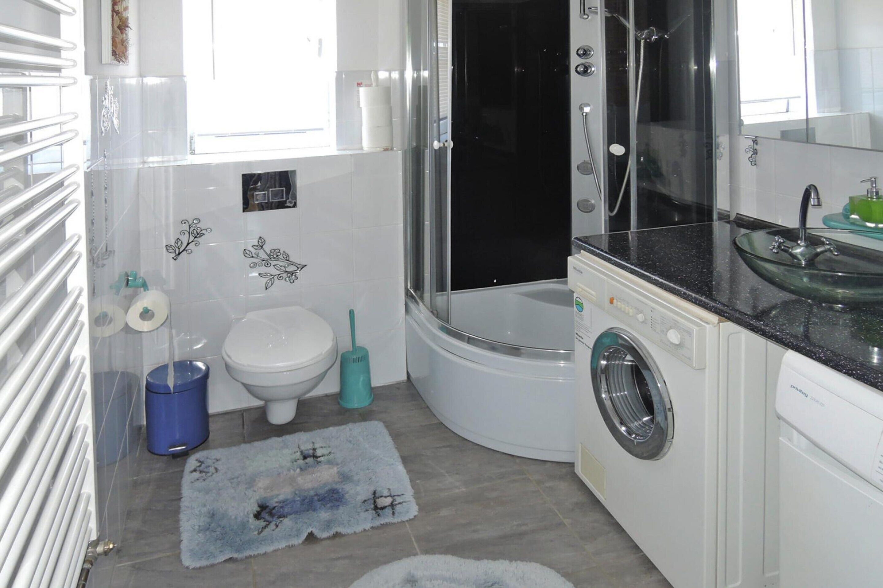 A compact bathroom with a glass-enclosed shower, a white toilet, a washing machine, and a sink with a glass basin.
