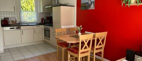 Une cuisine de taille réduite avec des meubles blancs, une cuisinière et un évier. On y trouve un coin repas avec une table et des chaises en bois. Un mur rouge orné d’un tableau et d’une plante est visible.