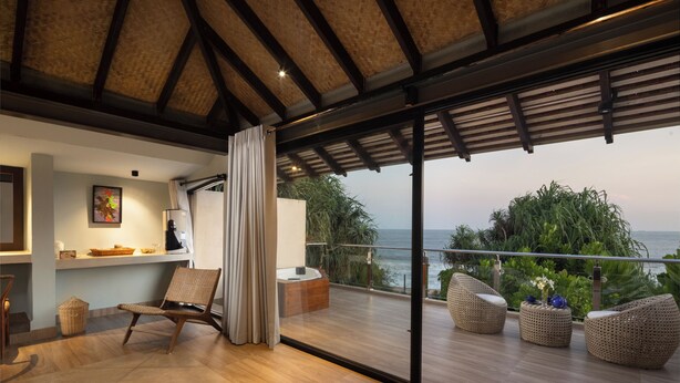 Un balcón moderno con piso de madera, una silla de mimbre, una mesita con un florero y vista al mar.