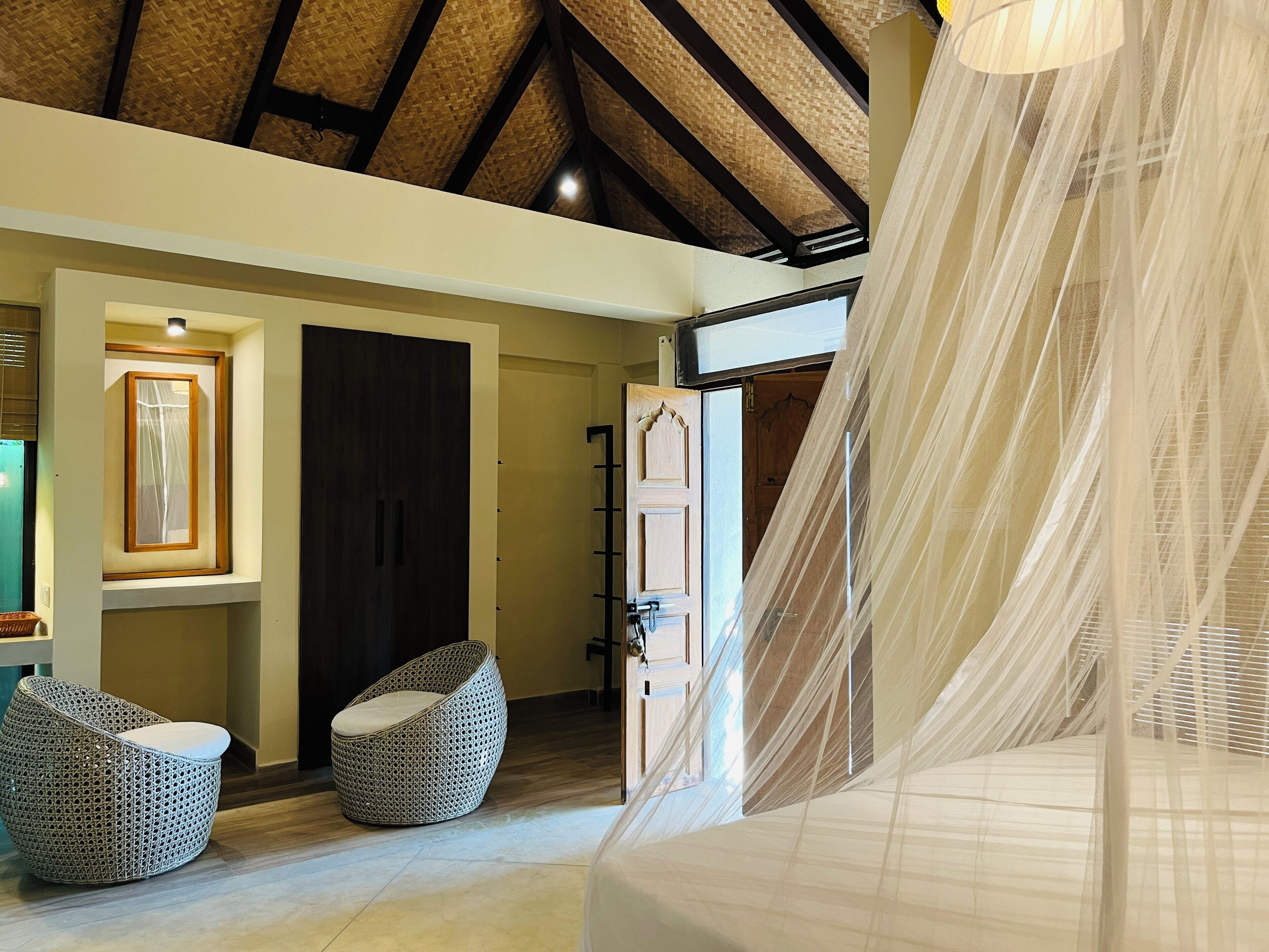 A room with a high ceiling, a bed with a canopy, two wicker chairs, a door, and a framed picture on the wall.