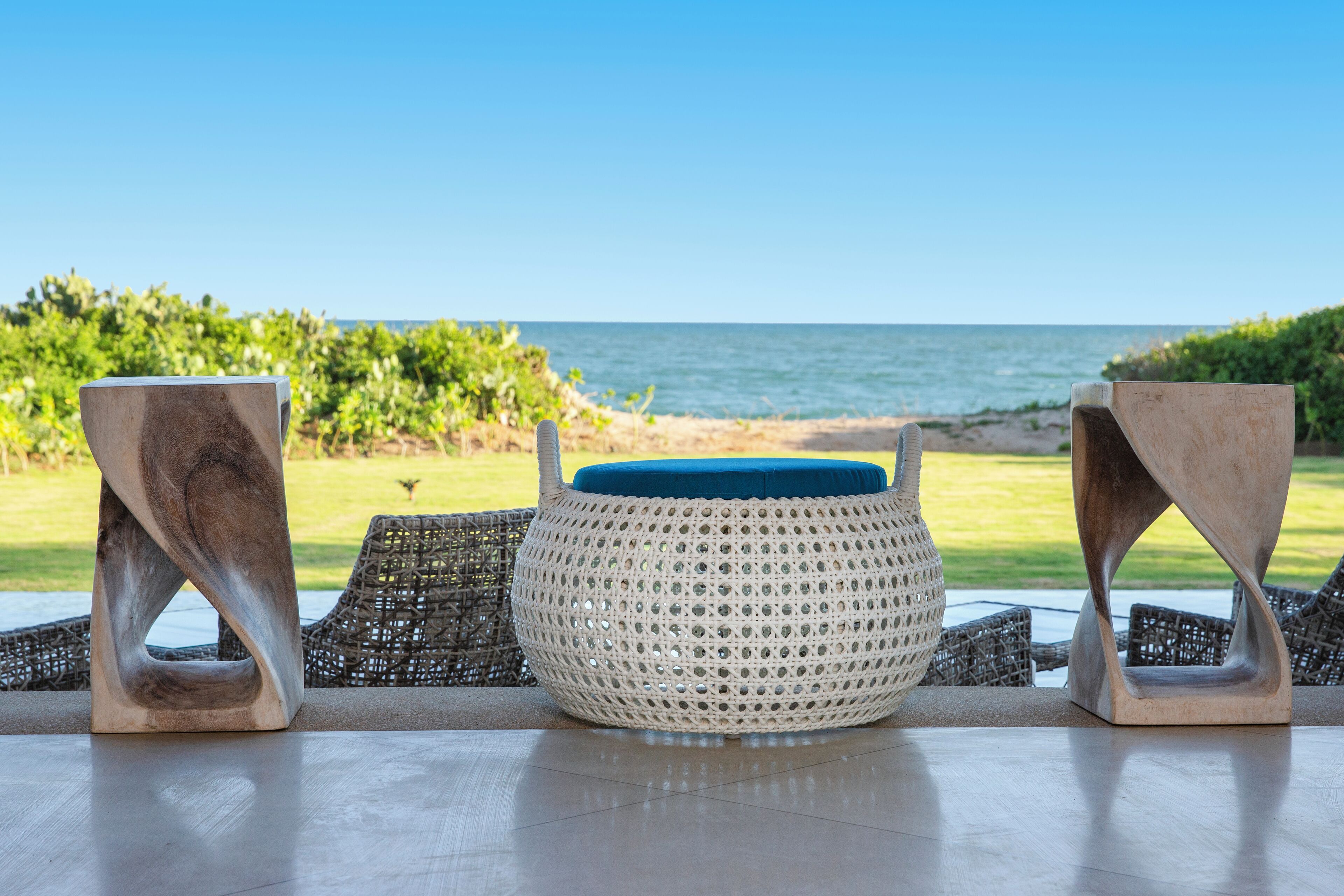 A wicker table with a woven basket centerpiece, two metal stools, and a view of the ocean.