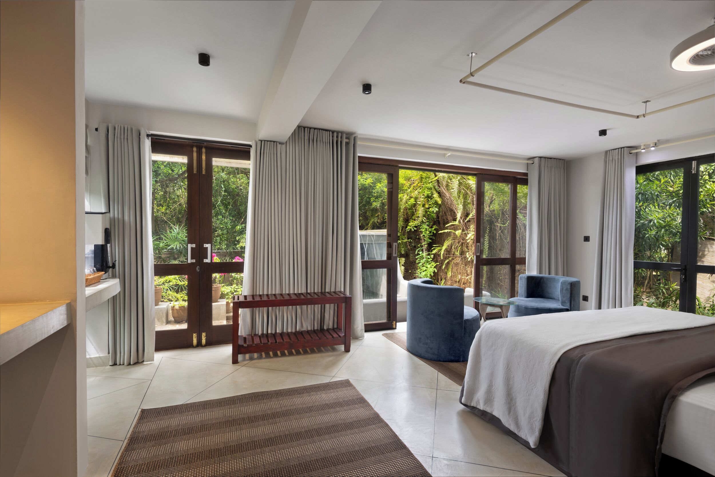 Una habitación de hotel moderna con una cama grande, una zona de estar con una silla azul, una mesita y vistas a vegetación exuberante a través de amplios ventanales.