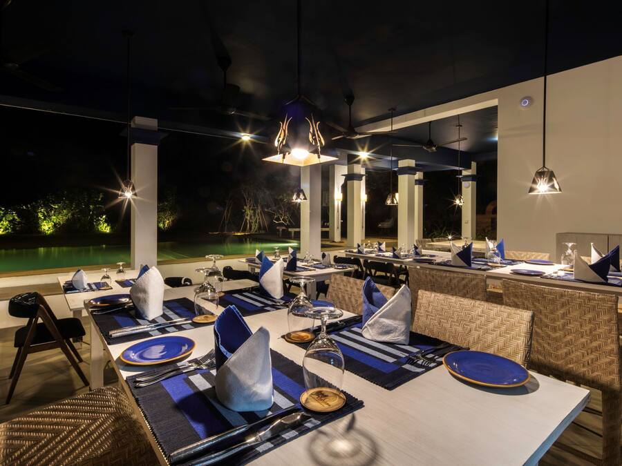 A restaurant with tables set for dining, a pool in the background, and hanging pendant lights.