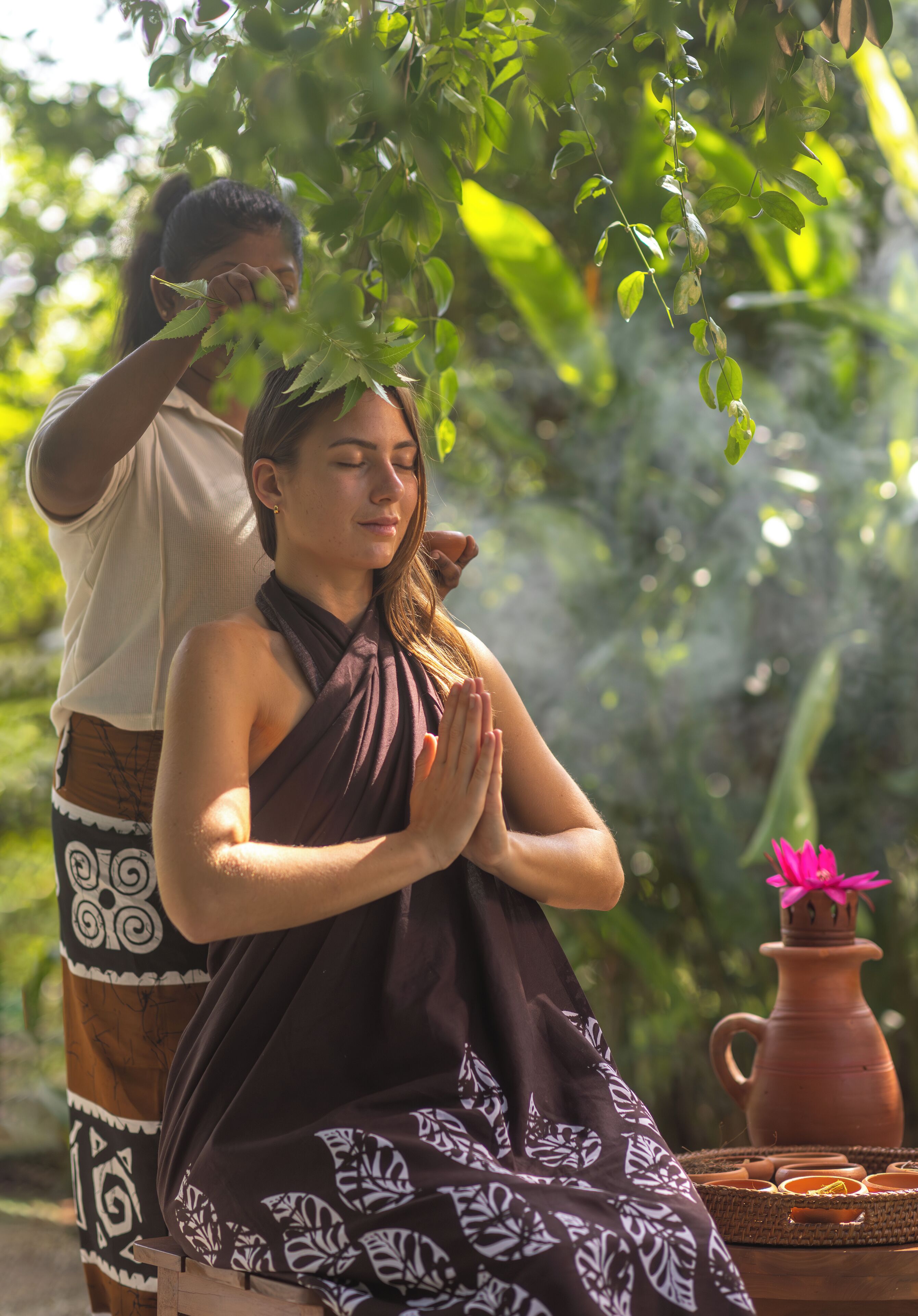 A person in a brown patterned dress sitting outdoors with a clay pot and a pink flower nearby.