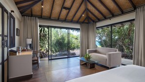 Amplia habitación con una cama grande, un sofá, una mesa de centro y un escritorio con una lámpara. Amplias ventanas ofrecen vistas a la vegetación exterior.