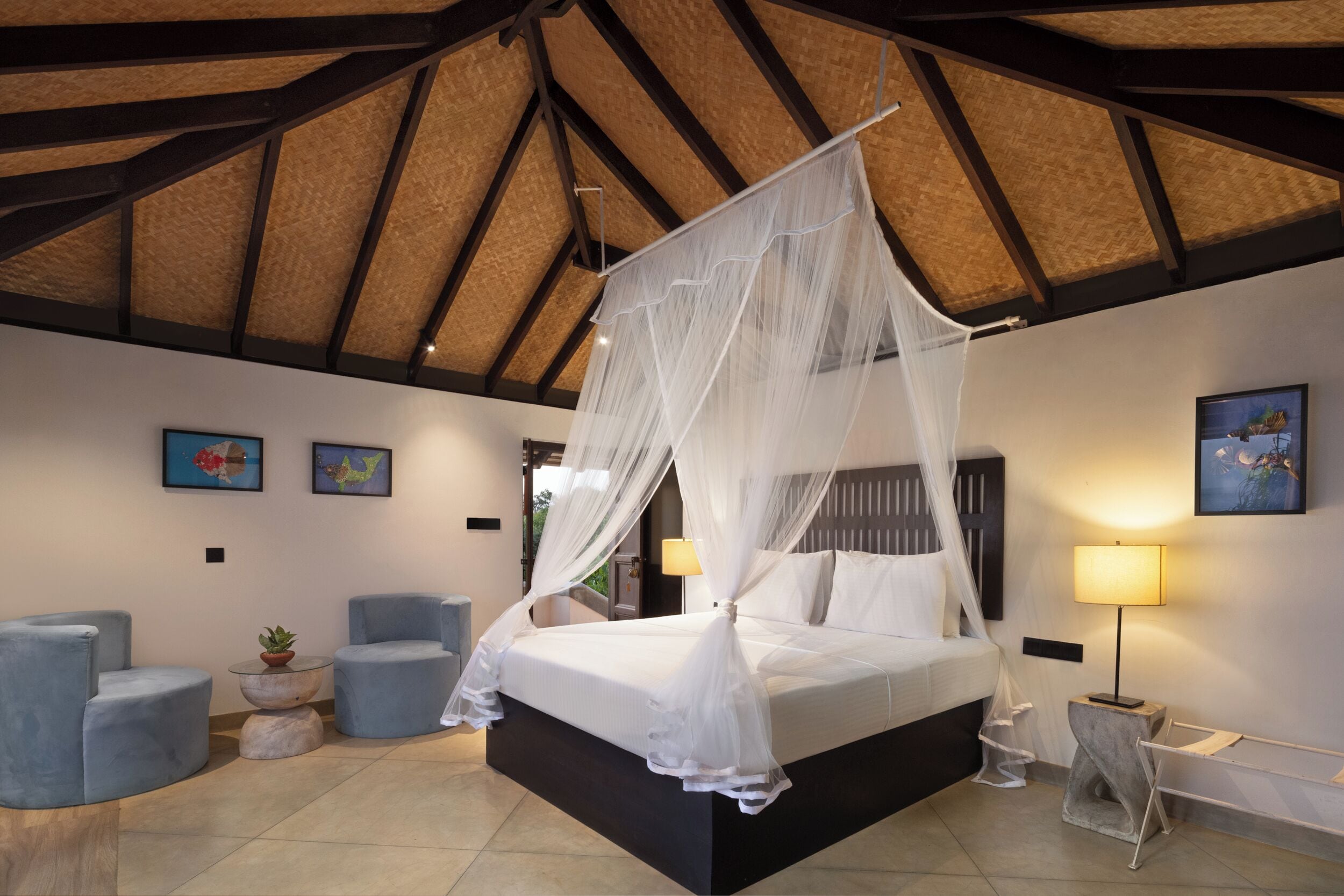 A bedroom with a bed under a canopy, two armchairs, a small table with a plant, and a nightstand with a lamp.