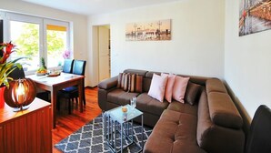 A modern living room with a brown sofa, a dining table with chairs, a coffee table, and a large window. - Comfortable 3-room Apartment Close to the sea (Ustronie Morskie)