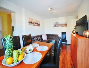 A modern living room with a dining area, featuring a wooden dining table with placemats, a sofa, and a television on a wooden cabinet. - Comfortable 3-room Apartment Close to the sea (Ustronie Morskie)