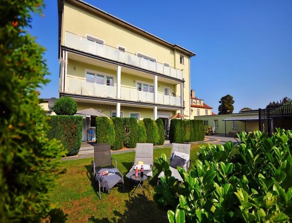 A two-story building with a balcony, a well-maintained garden with chairs and umbrellas, and a clear blue sky. - Comfortable 3-room Apartment Close to the sea (Ustronie Morskie)