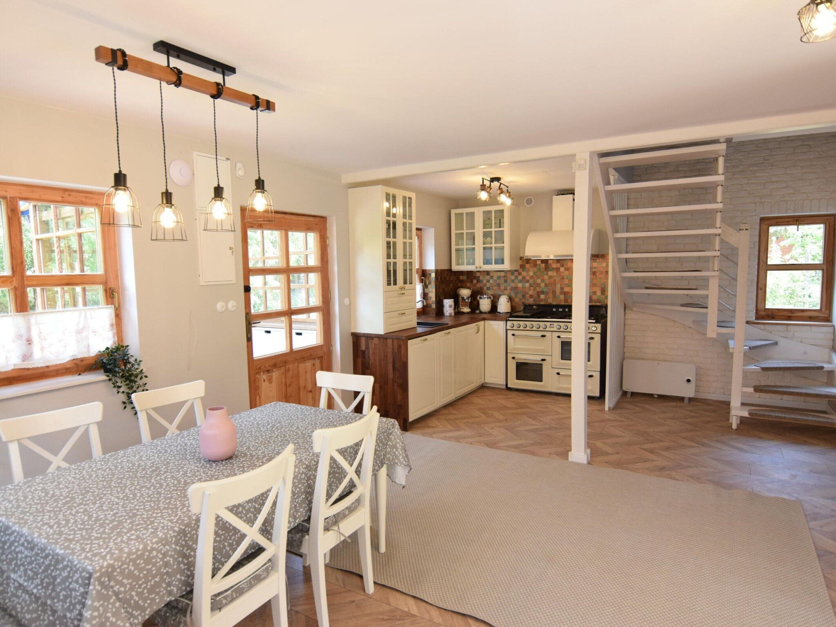 Une cuisine spacieuse avec des meubles blancs, un plan de travail en bois et un four encastré. On y trouve un coin repas avec une table et des chaises, ainsi qu’un escalier menant à l’étage supérieur.