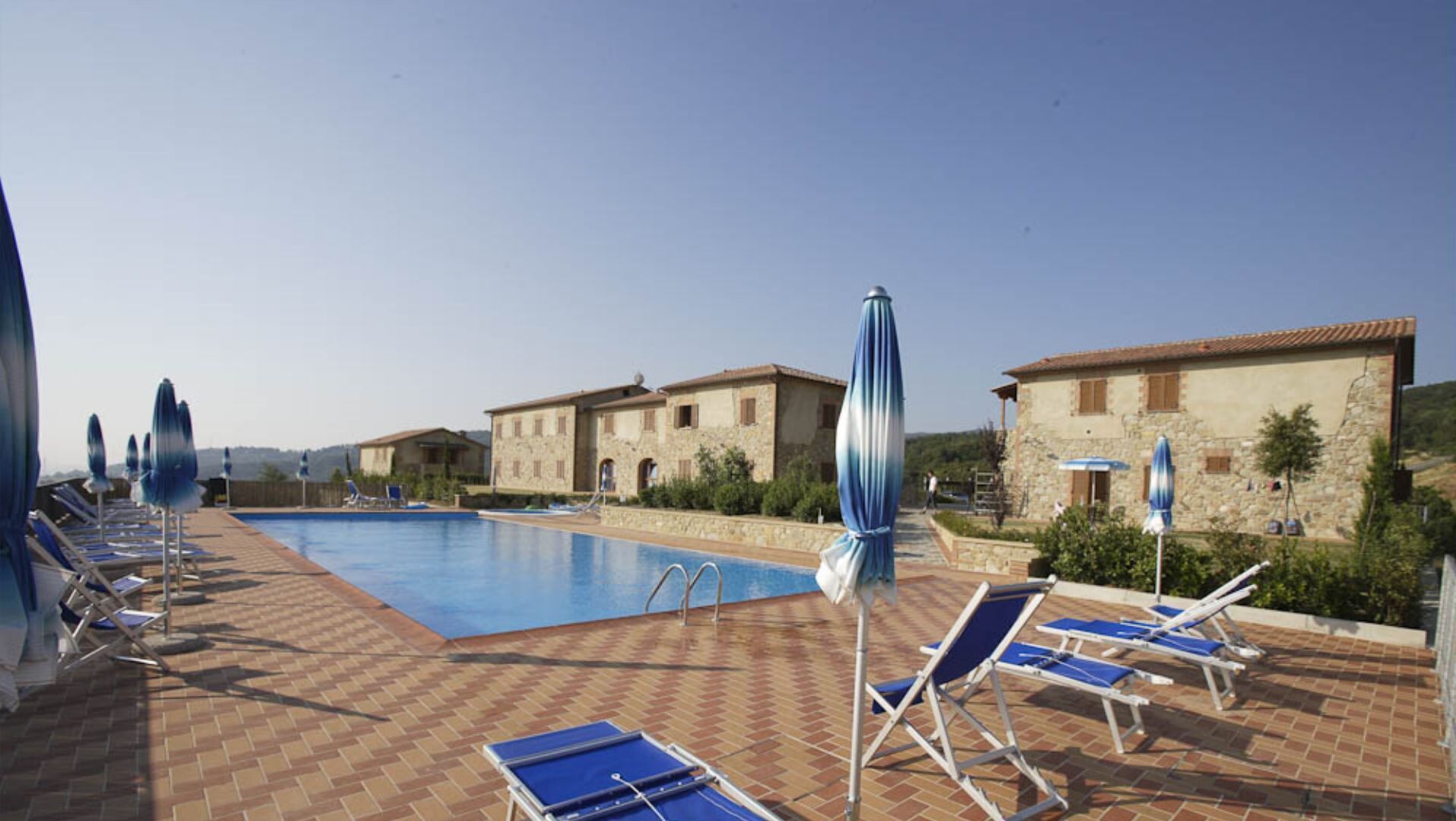 Un espace piscine avec des chaises longues et des parasols, des bâtiments en pierre et un ciel dégagé.