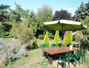 A wooden table with a green umbrella, surrounded by green chairs, set in a garden with various trees and shrubs. - Holiday Home Sulomino With Private Beach (gmina Wolin)