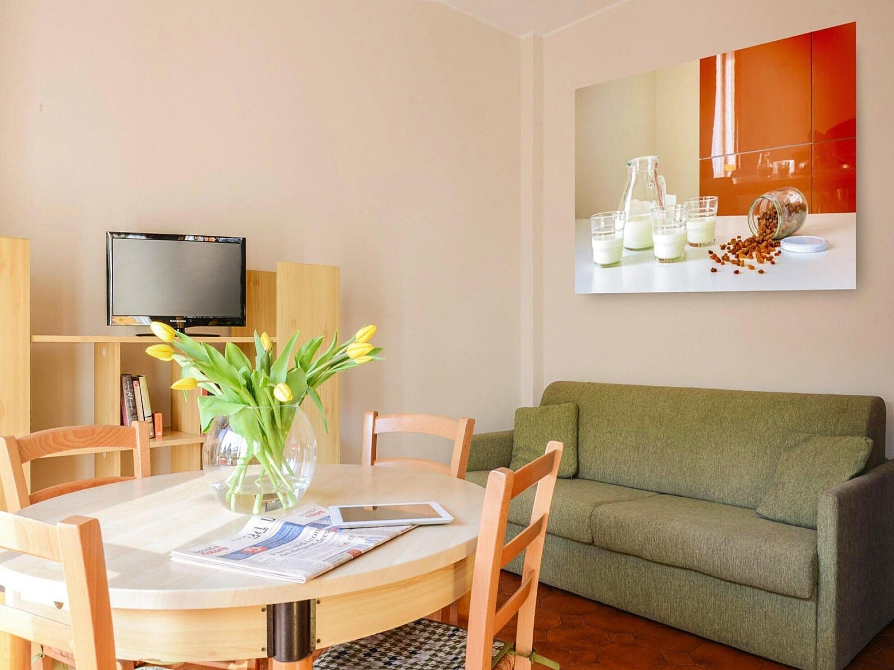 A modern living room with a round dining table, wooden chairs, a green sofa, a flat-screen TV, and a large painting on the wall.