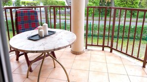 Une table ronde avec un plateau en marbre, une chaise à carreaux et un plateau avec des bougies, le tout sur un balcon donnant sur une clôture verte et des arbres.