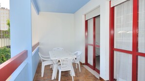 Un balcón con una mesa y sillas blancas, ventanas con marcos rojos y suelo de baldosas.