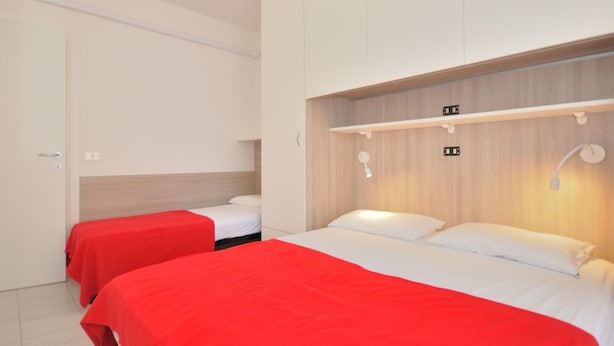 Habitación de hotel con dos camas, cada una con colcha roja, cabecero de madera y pared blanca. Hay dos mesitas de noche con lámparas.
