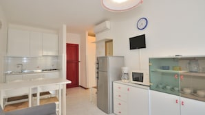 Una cocina compacta con armarios blancos, una puerta roja, una cafetera, un microondas y un reloj en la pared.