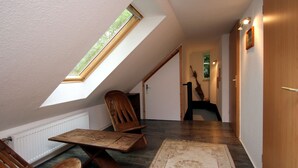 Ein schmaler Flur mit schrägverlaufender Decke, Holzsesseln, einem Tisch und einem Teppich mit Blumenmuster.