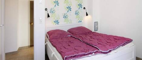 A single-bed room with a white headboard, a floral-patterned wall, two bedside lamps, and a red bedspread.