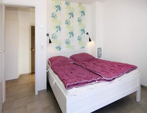 A single-bed room with a white headboard, a floral-patterned wall, two bedside lamps, and a red bedspread. - Ferienhaus, Boiensdorf (Boiensdorf)
