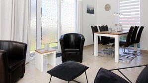Egy modern étkező, fehér asztallal és fekete székekkel, nagy ablakokkal, melyekre függöny van, és ahonnan zöld növényzetre nyílik kilátás.