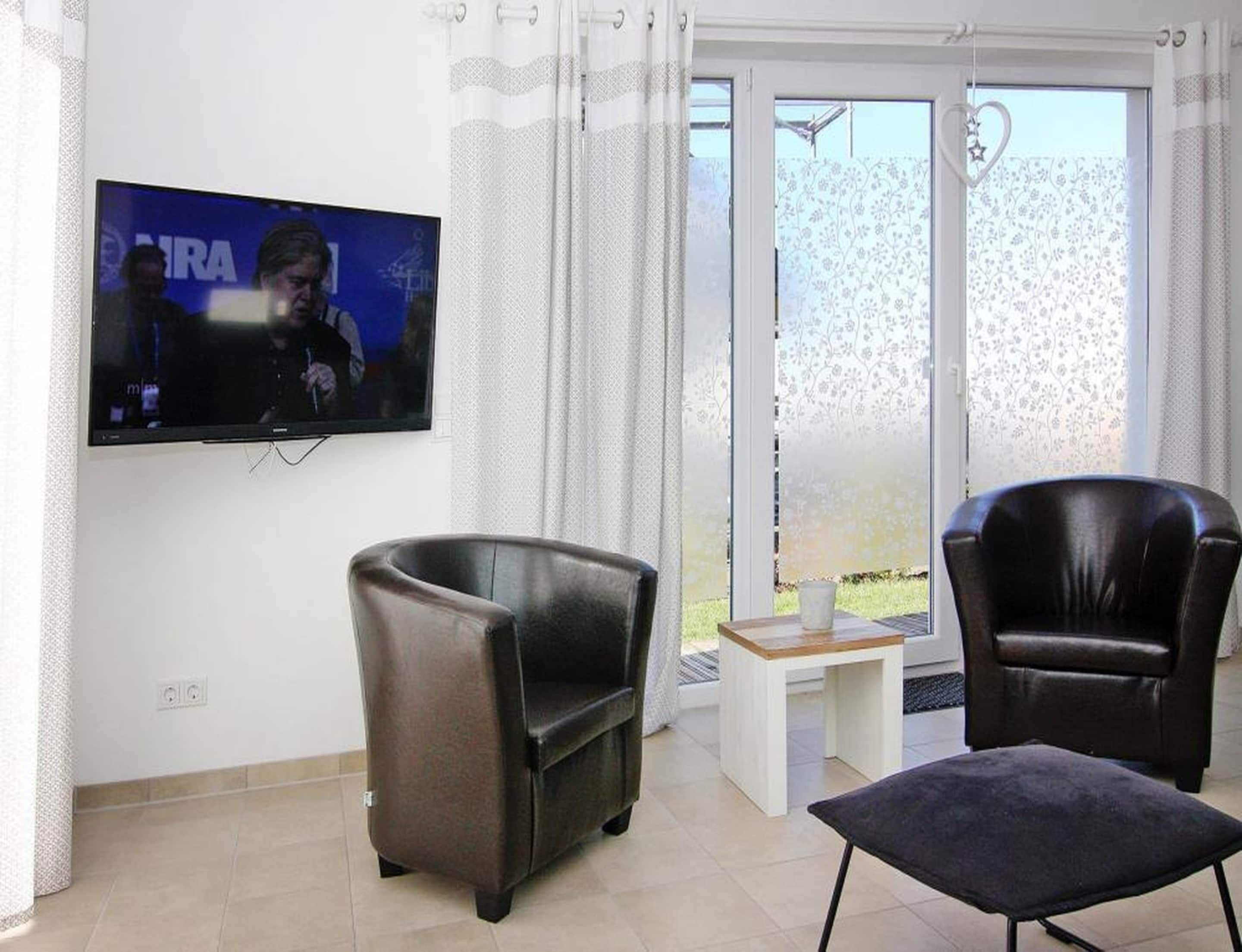 A living room with a flat-screen TV mounted on the wall, two leather armchairs, a small wooden table, and a sliding glass door leading to an outdoor area.