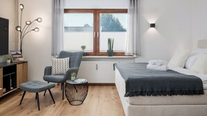 Een moderne slaapkamer met een bed, een stoel, een klein tafeltje, een televisie en een raam met gordijnen.