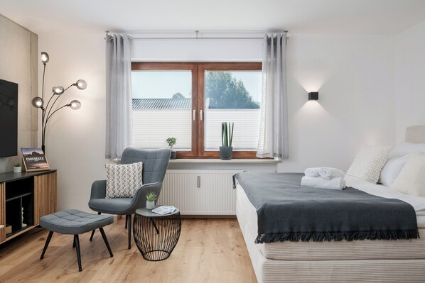 A modern bedroom with a bed, a chair, a small table, a TV, and a window with curtains.