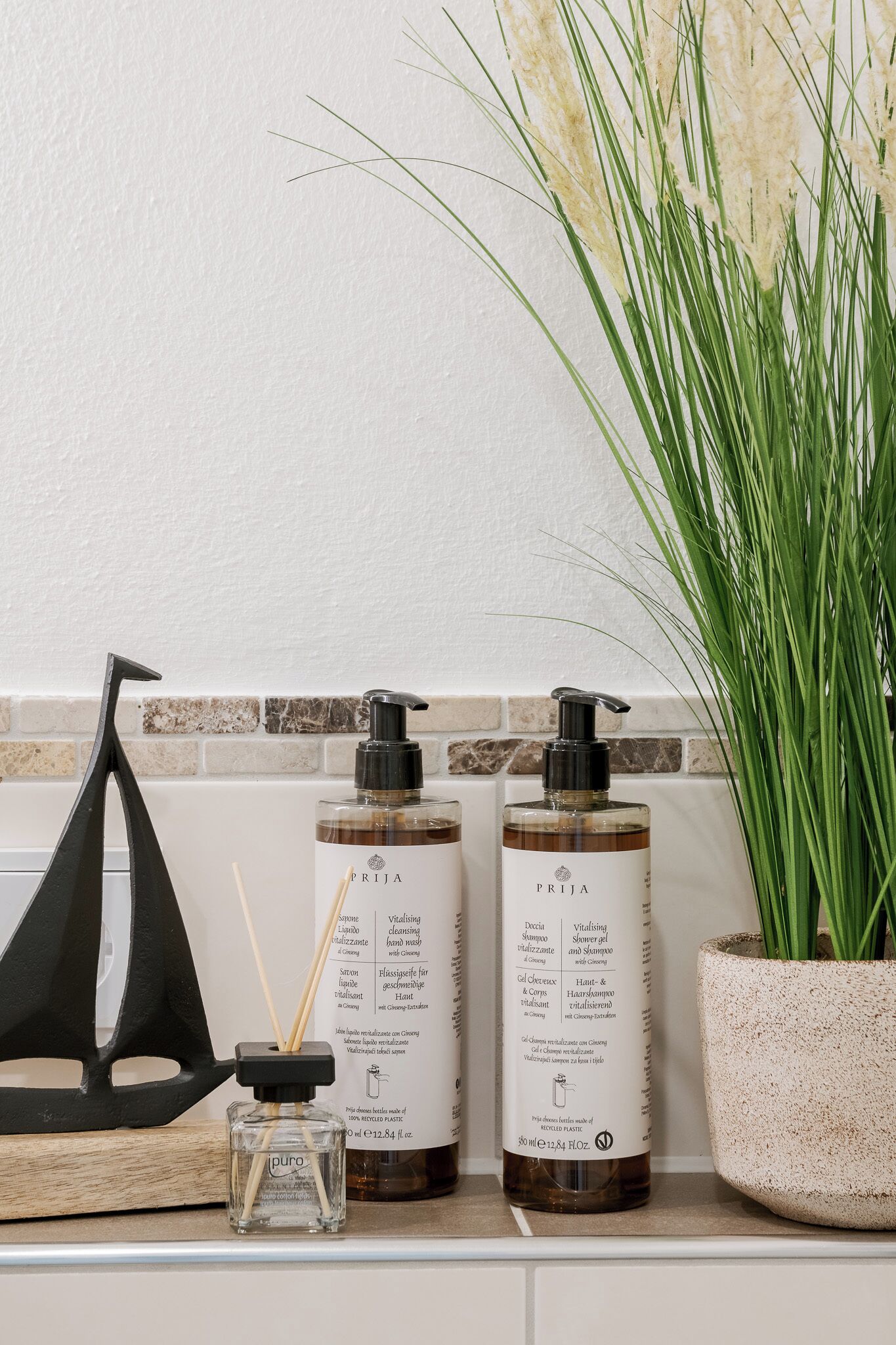 Dois frascos de produtos de cuidados para a pele PRIJA numa estante com uma planta em vaso e um objeto decorativo preto.