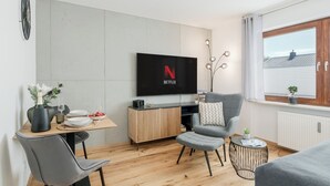 Een moderne woonkamer met een flatscreen-tv waarop het Netflix logo staat, een eethoek met een houten tafel en stoelen, en een gezellige zithoek met een grijze fauteuil en poef.