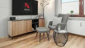 Een moderne woonkamer met een flatscreen televisie, een grijze fauteuil, een klein voetenbankje, een bijzettafeltje met een plant en een decoratieve lamp.