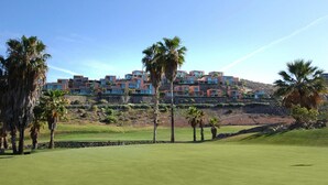 Um campo de golfe com edifícios modernos ao fundo, palmeiras e um céu limpo.