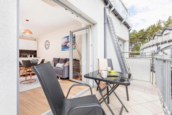 Une terrasse extérieure avec une table et des chaises, une porte-fenêtre coulissante donnant sur une cuisine et un mur extérieur blanc.