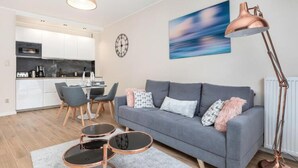 A modern living room with a grey sofa, two round coffee tables, a dining area with chairs, a kitchen in the background, and a large abstract painting on the wall. - Apartment With Swimming Pool, Terrace, Pobierowo (Rewal)