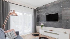 A modern living room with a grey sofa, a white entertainment unit, a flat-screen TV mounted on a grey stone wall, and a large window with grey curtains. - Apartment With Swimming Pool, Terrace, Pobierowo (Rewal)