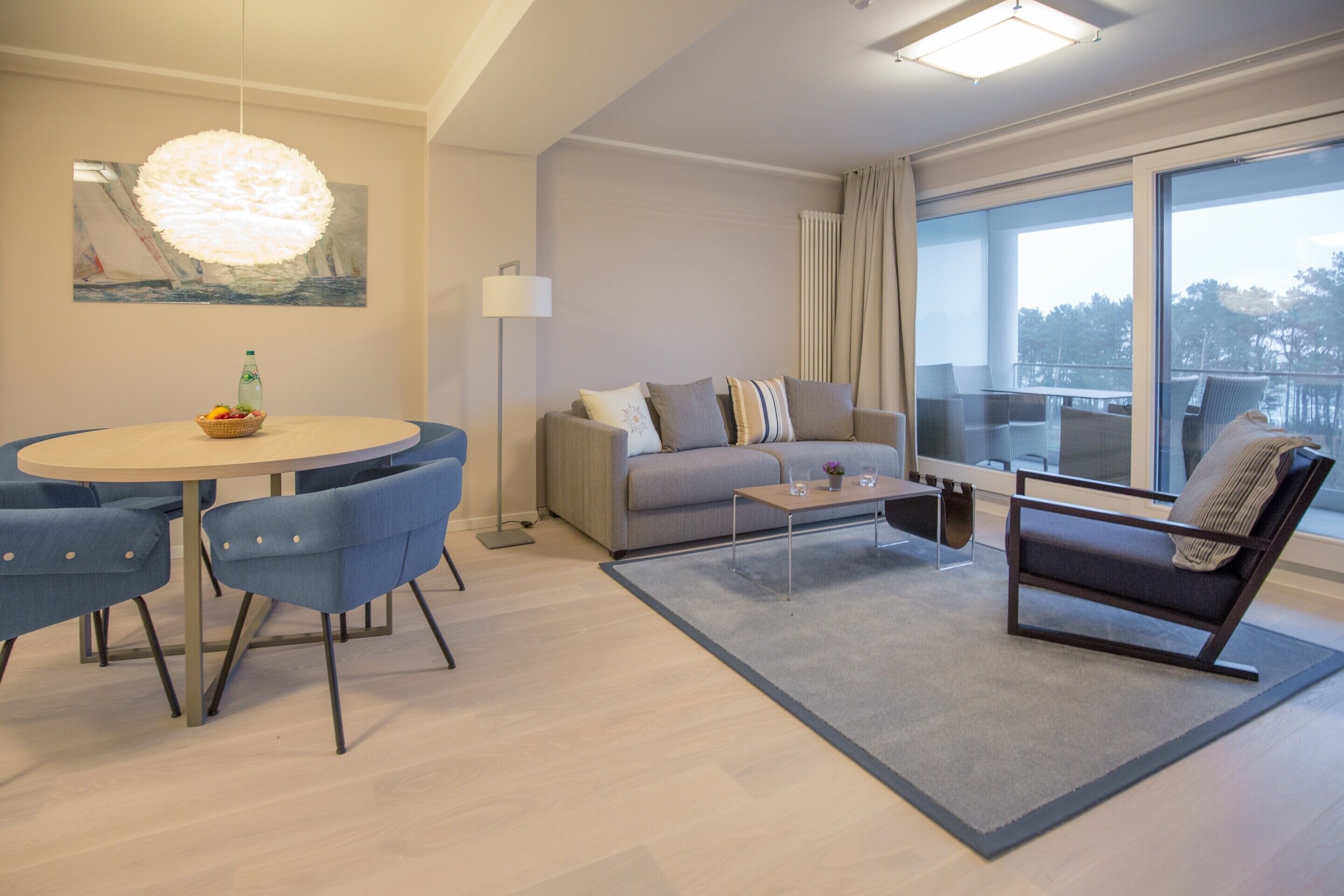 A modern living room with a round dining table, blue chairs, a grey sofa, a black armchair, and a large window with a view.