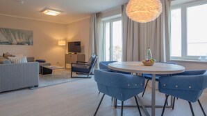 A modern living room with a round dining table, blue chairs, a sofa, a TV, and a large window with curtains. - Dunenresidenz Prora (Binz)