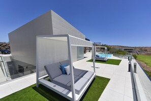 A modern outdoor seating area with a white canopy bed and a pool in the background.