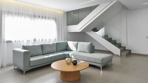 Una sala de estar moderna con un sofá seccional gris claro, una mesa de centro de madera redonda y una escalera con barandillas de vidrio.