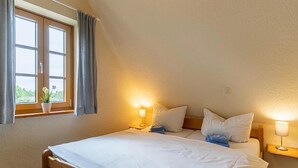 A bedroom with a wooden bed, two bedside lamps, a window with curtains, and a view of greenery outside. - Landhaus in Vollerwiek (Vollerwiek)