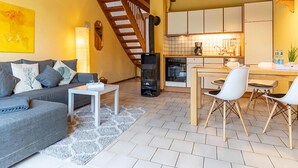 A cozy living room with a sofa, coffee table, and a fireplace. A kitchen with white cabinets and a dining area with chairs is visible in the background. - Landhaus in Vollerwiek (Vollerwiek)
