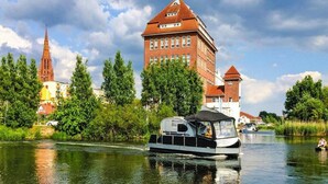 A boat on a river with a red brick building and a church in the background. - Hausboot auf der Peene, Demmin (Demmin)