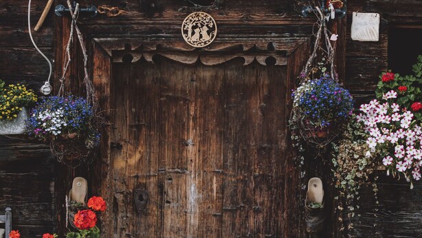 A rustic wooden door with a decorative emblem, surrounded by hanging flower baskets and vibrant flowers. - Apartment in Fusch Near Grossglockner Road (Fusch an der Grossglocknerstrasse)