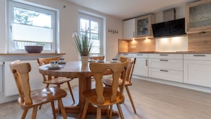 A modern kitchen with white cabinetry, a wooden dining table, and chairs. There are windows with blinds, a plant on the windowsill, and a bowl on the window sill. - Ferienwohnung fur Familien in St.peter Ording (St. Peter-Ording)