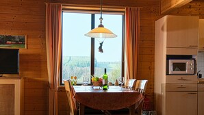 A wooden interior with a dining table, chairs, a window with curtains, a hanging light fixture, a television, and a microwave. - Doppelhaushalfte in Hasselfelde (Oberharz am Brocken)