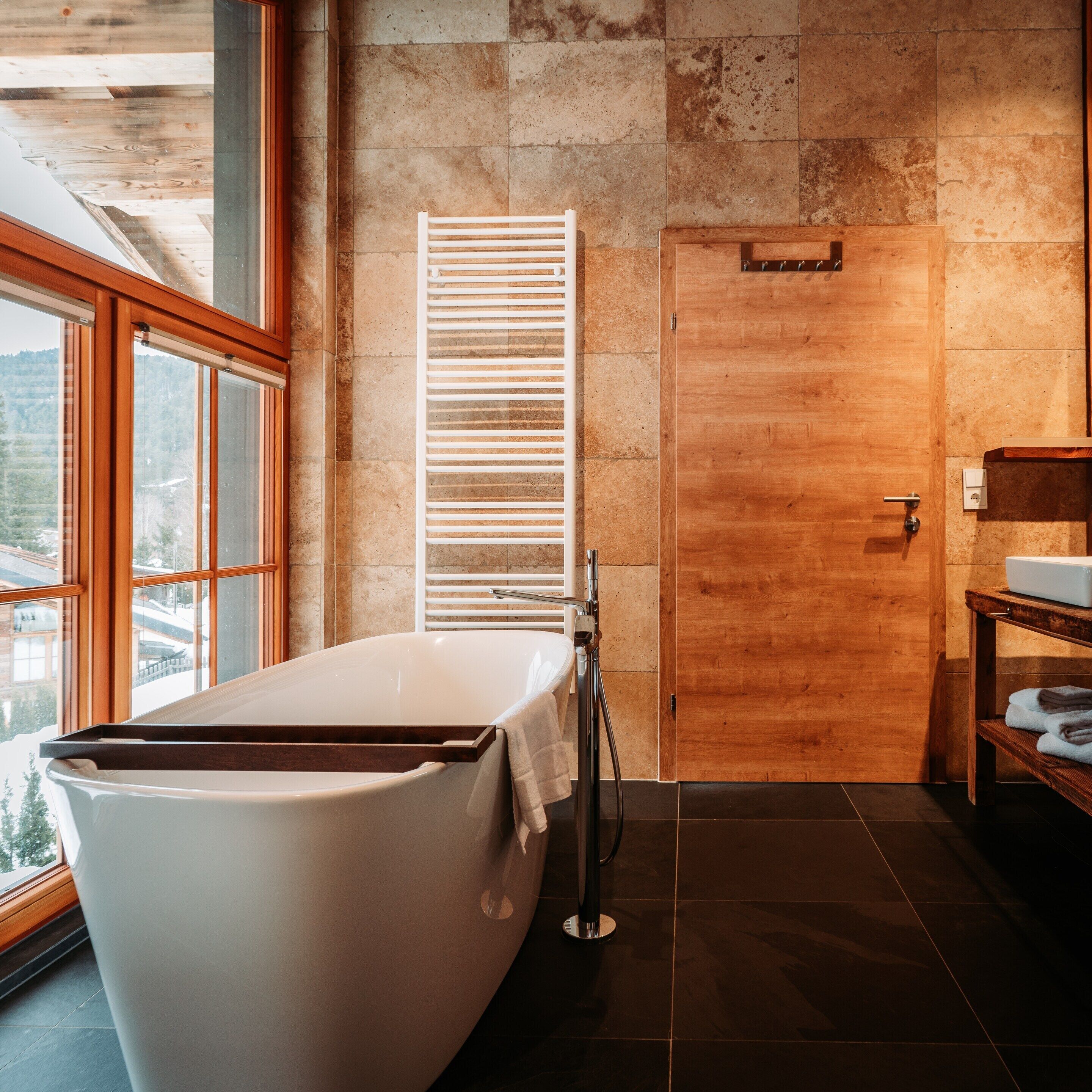 A modern bathroom with a freestanding bathtub, a wooden door, and a window with a view of the outdoors.