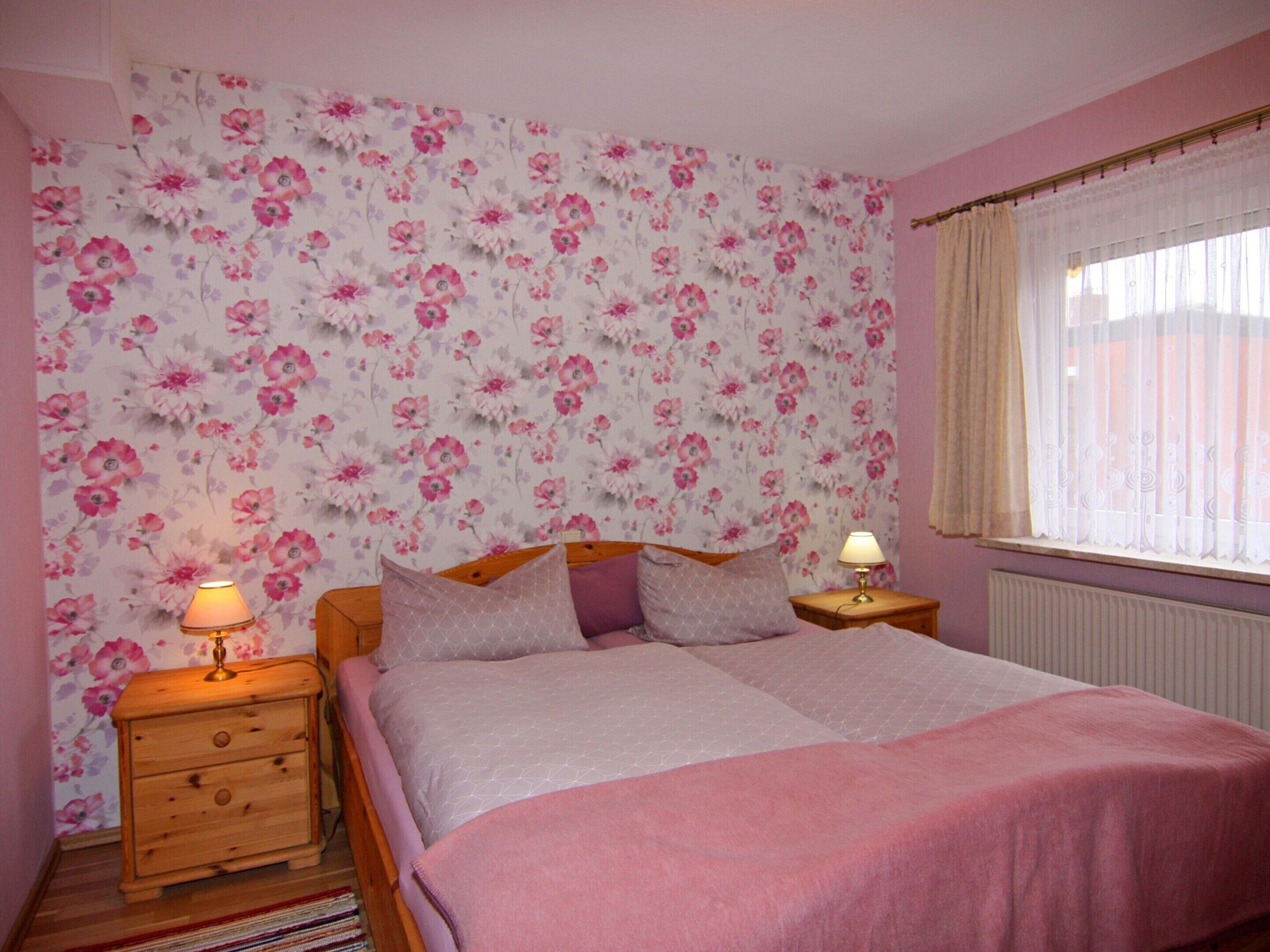 Un dormitorio con papel tapiz floral, una cama con manta rosa, dos mesitas de noche con lámparas y una ventana con cortinas.