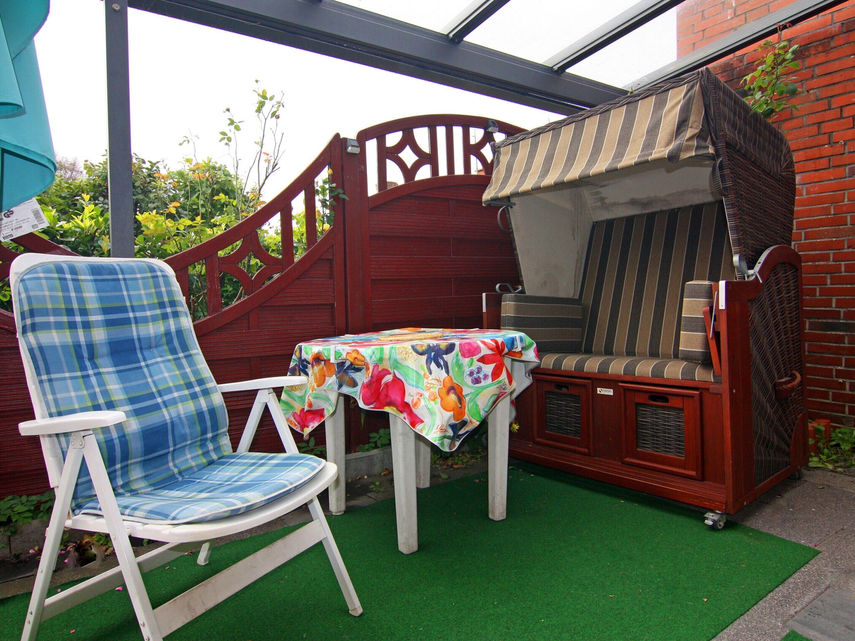Un espace patio avec une chaise pliante blanche, une table recouverte d’une nappe à motifs floraux et un banc en bois agrémenté d’un coussin à rayures.